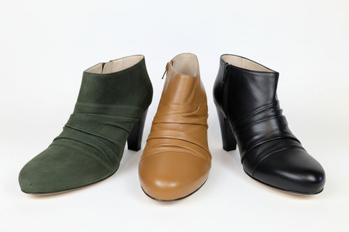 Cassidy women's ankle boots with block heels are displayed side-by-side on a white background. From left to right, the boots are dark green suede, Cognac leather, and black leather, each featuring a unique gathered, pleated design on the front. Zipper closure, runs narrow. We recommend going up by 1/2 size.