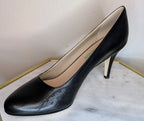 Kate black nappa leather high-heeled pump with a pointed toe and subtle perforated detailing on the vamp, displayed on a light marble surface with a gold-framed edge against a neutral wall.