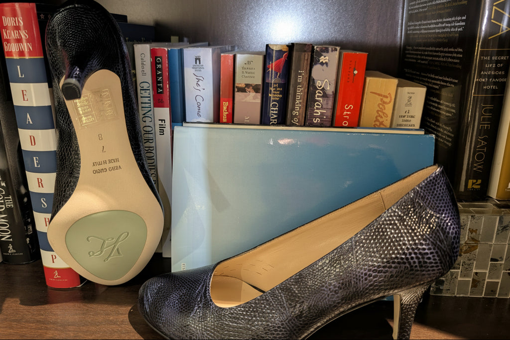Kate women's high-heeled pumps with a Navy textured python-like snakeskin pattern, displayed with side  and bottom views on a wooden bookshelf alongside a light blue shoebox and books.