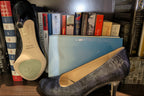 Kate women's high-heeled pumps with a Navy textured python-like snakeskin pattern, displayed with side  and bottom views on a wooden bookshelf alongside a light blue shoebox and books.
