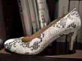 JL Shoes - Salem women's high-heeled leather pumps with a black and white snakeskin pattern, resting on a dark wooden shelf in front of a bookshelf background.